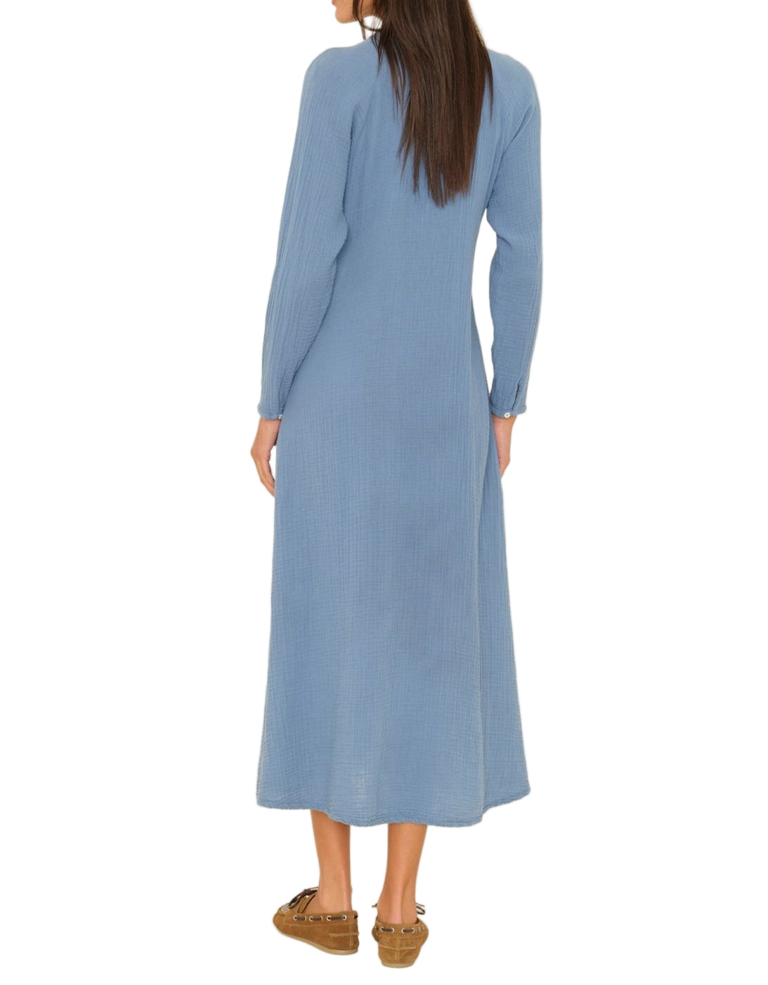 A woman is shown from the back wearing the Xirena Tabitha Dress, a flowy midi dress with long sleeves, paired with blue and brown moccasin-style shoes.