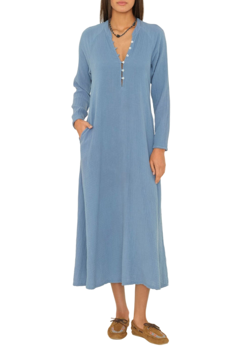 A woman wearing the Xirena Tabitha Dress, a light blue, long-sleeve cotton gauze dress with a buttoned neckline, and brown flat shoes.