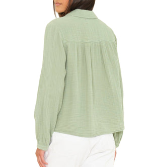 A woman is seen from the back wearing the Xirena Julianne Shirt, a light green collared, long-sleeve button-front top, paired with white pants against a plain background.