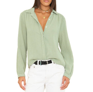 A woman stands against a white background wearing the Xirena Julianne Shirt in light green cotton gauze, tucked into white jeans with a black belt.