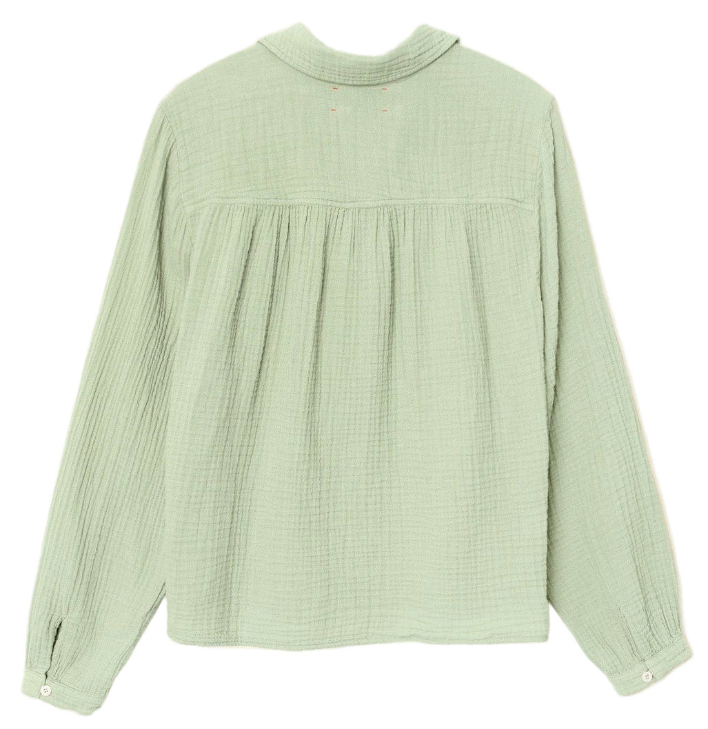 The Julianne Shirt by Xirena is a light green, button-front top in textured double-faced cotton gauze, featuring buttoned cuffs and a gathered back yoke, laid flat to highlight its relaxed style.