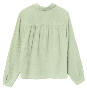The Julianne Shirt by Xirena is a light green, button-front top in textured double-faced cotton gauze, featuring buttoned cuffs and a gathered back yoke, laid flat to highlight its relaxed style.