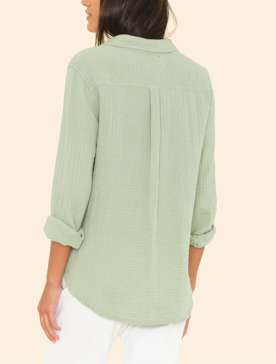A woman with long dark hair faces away, modeling the Xirena Scout Shirt in light green cotton gauze with rolled sleeves and white pants against a plain beige background.