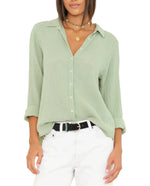 A woman wears the Xirena Scout Shirt in light green with rolled sleeves, paired with white pants, a black belt, and layered gold necklaces.