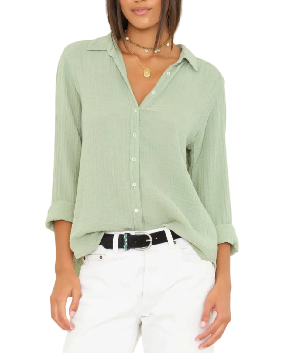 A woman wears the Xirena Scout Shirt in light green with rolled sleeves, paired with white pants, a black belt, and layered gold necklaces.