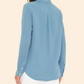 A woman faces away, wearing the Xirena Scout Shirt in soft crinkled cotton gauze light blue, paired with white pants, against a plain beige background.