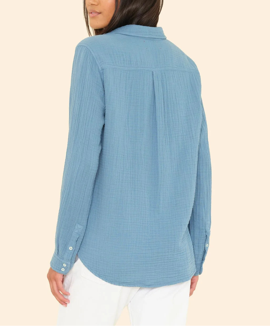 A woman faces away, wearing the Xirena Scout Shirt in soft crinkled cotton gauze light blue, paired with white pants, against a plain beige background.