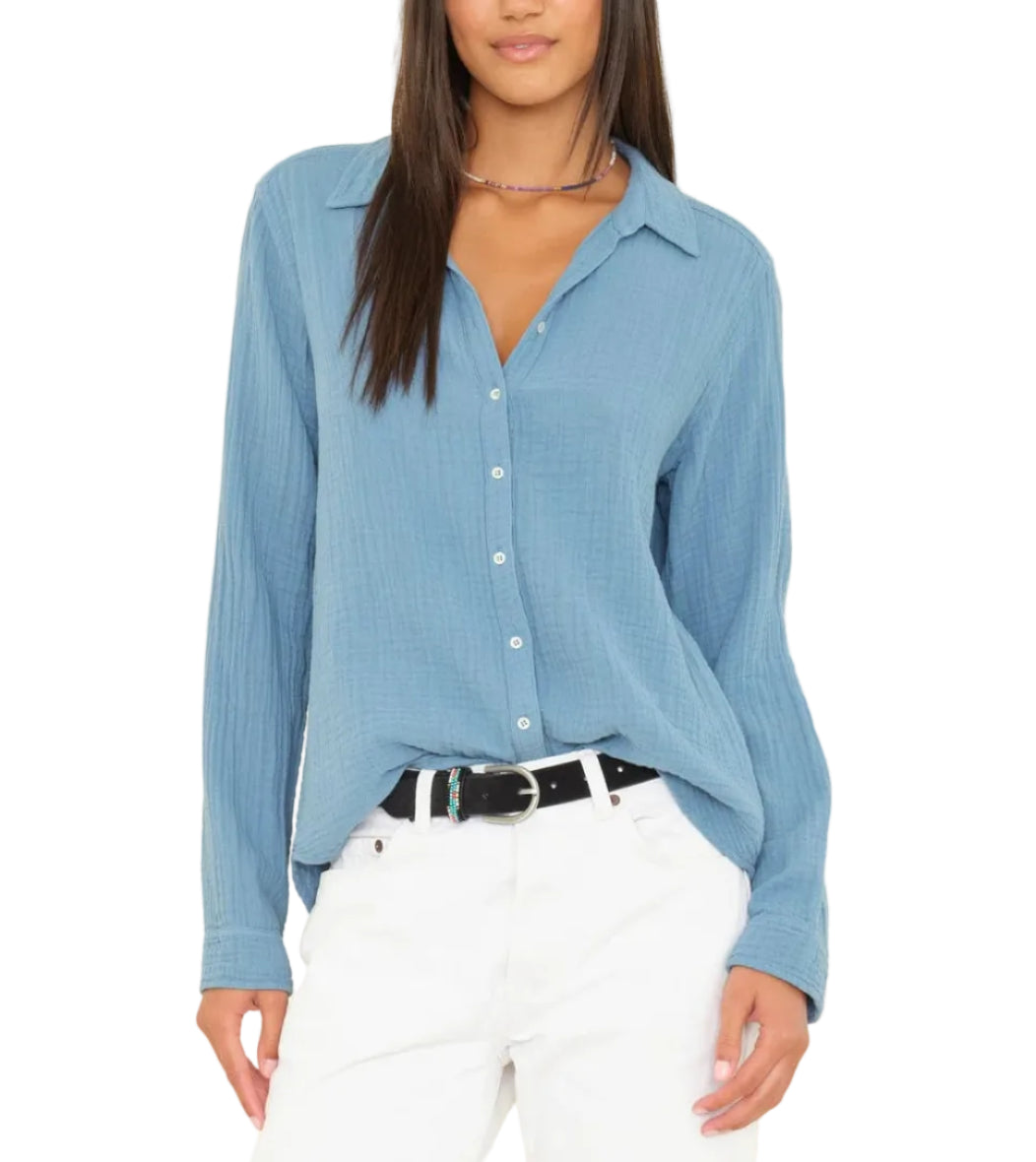 A person wearing the Xirena Scout Shirt in light blue, paired with white pants and a black belt, stands with one hand in their pocket.