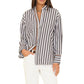 Wearing the Xirena Gillian Shirt, a relaxed button-front style with black and white vertical stripes, and white pants, a person stands against a plain white background with one hand in their pocket.