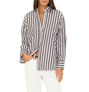 Wearing the Xirena Gillian Shirt, a relaxed button-front style with black and white vertical stripes, and white pants, a person stands against a plain white background with one hand in their pocket.