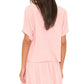 A person with long brown hair is seen from the back, wearing the Xirena Carson Top in light pink cotton gauze with a matching skirt, set against a white background.