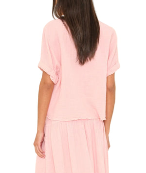 A person with long brown hair is seen from the back, wearing the Xirena Carson Top in light pink cotton gauze with a matching skirt, set against a white background.