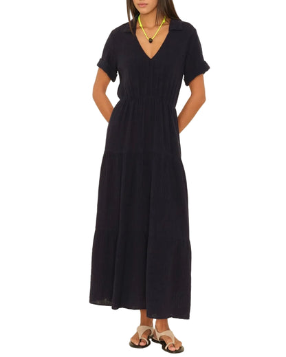 A woman wears the black Larissa Dress by Xirena featuring cotton gauze, short sleeves, a V-neck, elastic waist, and layered skirt. Styled with sandals and a necklace, her hands rest behind her back.