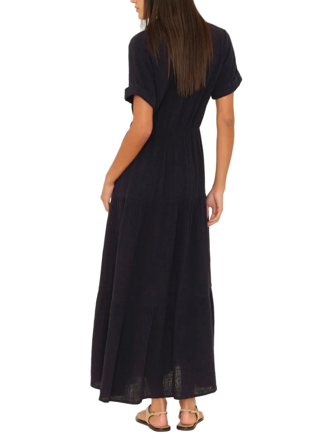 A woman faces away, wearing the Xirena Larissa Dress—a black cotton gauze style with an elastic waist—paired with flat sandals against a white background.