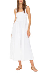 A woman wearing the Xirena Florence Dress, a sleeveless white maxi dress with a smocked bodice, paired with tan flat sandals and standing against a white background.