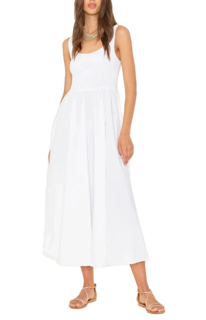 A woman wearing the Xirena Florence Dress, a sleeveless white maxi dress with a smocked bodice, paired with tan flat sandals and standing against a white background.