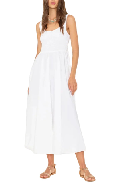 A woman wearing the Xirena Florence Dress, a sleeveless white maxi dress with a smocked bodice, paired with tan flat sandals and standing against a white background.