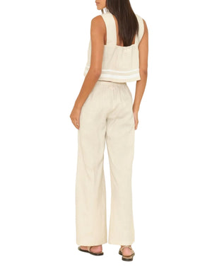A woman with her hair down stands barefoot, wearing Xirena’s Pippen Linen Pant—high-waisted, wide leg, and beige—paired with a matching sleeveless crop top with wide straps.