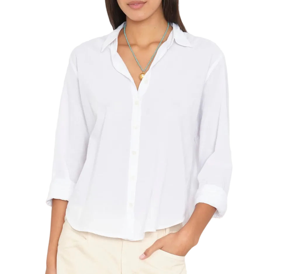 A woman stands with one hand in her pocket, wearing the Xirena Bowie Shirt—a modern silhouette white cotton poplin shirt with rolled-up sleeves—paired with beige pants and two necklaces.