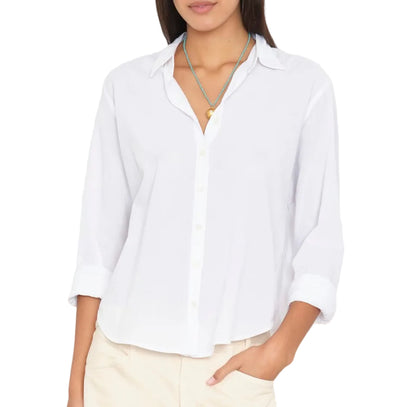 A woman stands with one hand in her pocket, wearing the Xirena Bowie Shirt—a modern silhouette white cotton poplin shirt with rolled-up sleeves—paired with beige pants and two necklaces.