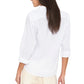 A woman is seen from behind wearing the long-sleeved white Bowie Shirt by Xirena, tailored from lightweight cotton poplin and paired with light-colored pants. Her sleeves are rolled up, and her straight brown hair is worn down, highlighting the shirt’s modern silhouette against a white background.