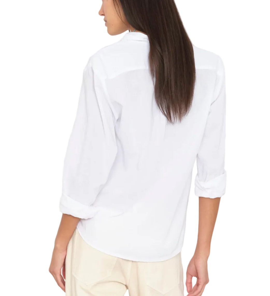 A woman is seen from behind wearing the long-sleeved white Bowie Shirt by Xirena, tailored from lightweight cotton poplin and paired with light-colored pants. Her sleeves are rolled up, and her straight brown hair is worn down, highlighting the shirt’s modern silhouette against a white background.