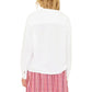 A person with wavy blonde hair is shown from the back, wearing the Xirena Cara Top, a white long-sleeve blouse, paired with a pink plaid skirt.