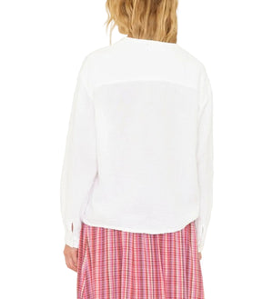 A person with wavy blonde hair is shown from the back, wearing the Xirena Cara Top, a white long-sleeve blouse, paired with a pink plaid skirt.