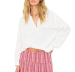 A woman wears the Xirena Cara Top, a loose white henley blouse with raglan sleeves, paired with a pink plaid tiered skirt, brown belt, and pink necklace. She poses with one hand on her hip against a white background.