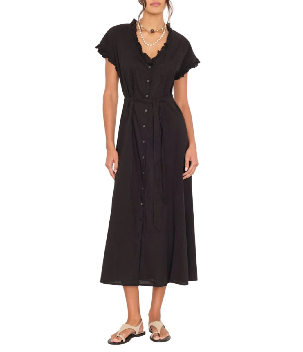 A woman wears the Xirena Dixie Dress, a black cotton poplin button-front midi with a tie waist, styled with sandals and a layered necklace, standing against a white background.