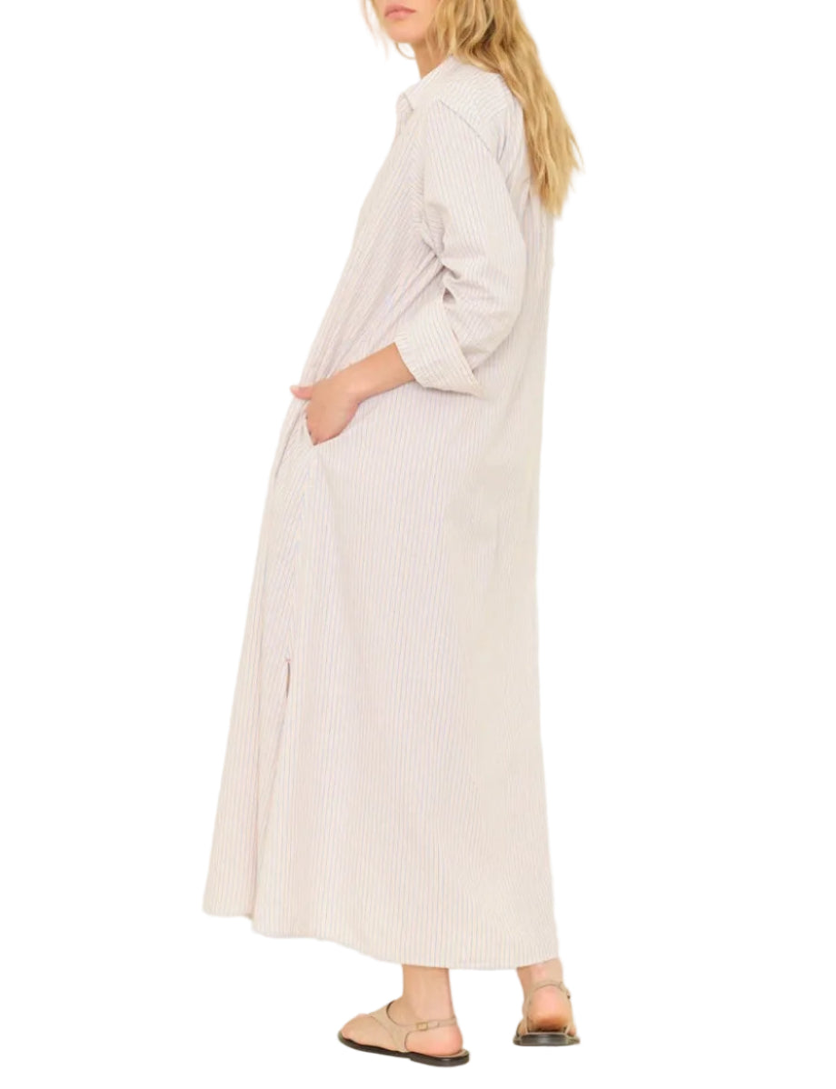A woman in the Xirena Boden Stripe Dress—a long, lightweight cotton shirtdress with stripes, long sleeves, side pockets, and button front—paired with flat sandals, stands sideways.