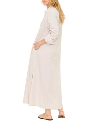 A woman in the Xirena Boden Stripe Dress—a long, lightweight cotton shirtdress with stripes, long sleeves, side pockets, and button front—paired with flat sandals, stands sideways.