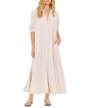 A person wears the Xirena Boden Stripe Dress, a long, lightweight cotton button-front shirtdress with rolled-up sleeves and side slits, paired with flat sandals.