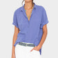 A woman wears the Xirena Channing Cotton Poplin Shirt with rolled sleeves, layered necklaces, white pants, and a patterned belt, standing against a plain background.