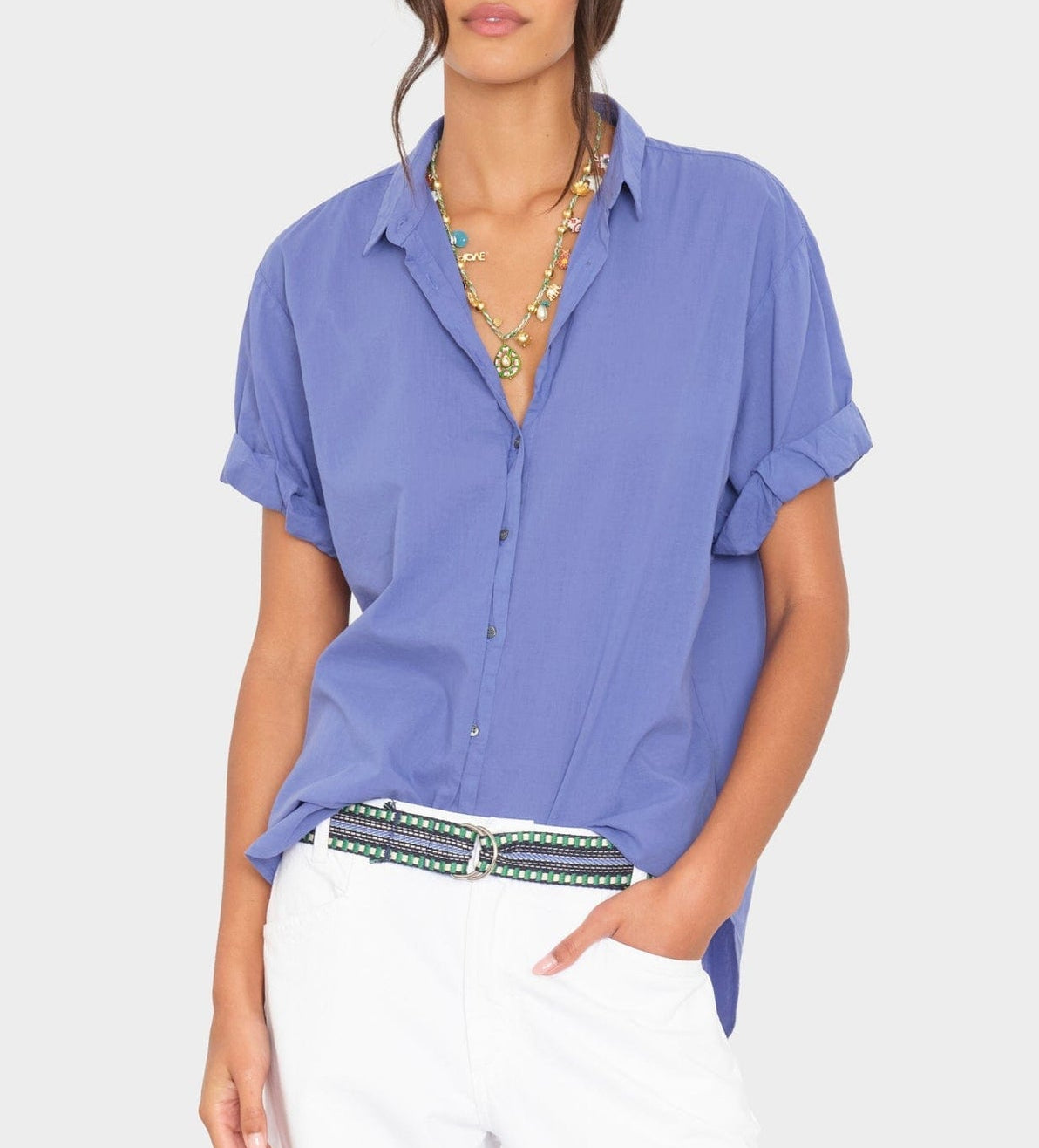 A woman wears the Xirena Channing Cotton Poplin Shirt with rolled sleeves, layered necklaces, white pants, and a patterned belt, standing against a plain background.