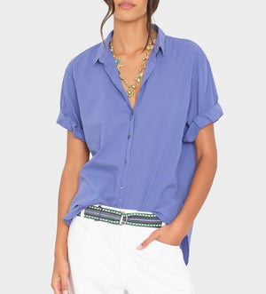 A woman wears the Xirena Channing Cotton Poplin Shirt with rolled sleeves, layered necklaces, white pants, and a patterned belt, standing against a plain background.