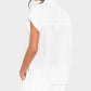 A woman is shown from the back wearing the Xirena Lexi Shirt, a loose-fitting, lightweight cotton short-sleeve top, paired with white pants against a light gray background.