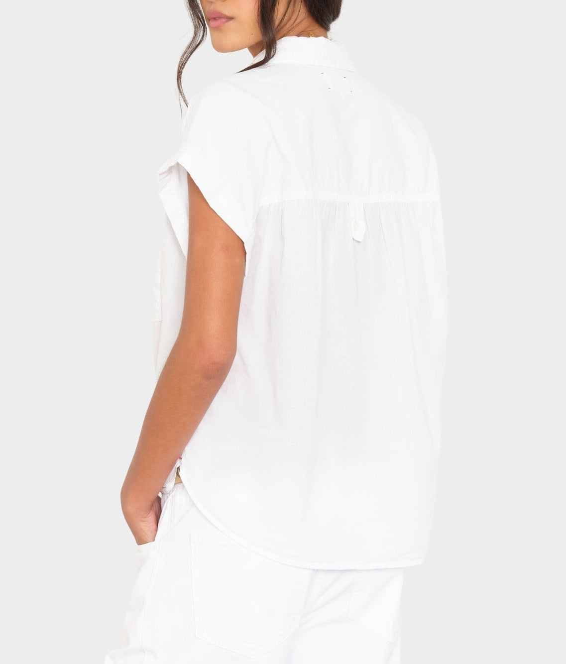 A woman is shown from the back wearing the Xirena Lexi Shirt, a loose-fitting, lightweight cotton short-sleeve top, paired with white pants against a light gray background.