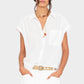 Wearing the Xirena Lexi Shirt, white pants, a beige belt, and layered necklaces, a person stands with hands in pockets.