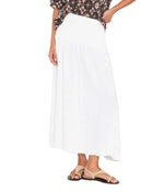 A person stands with one hand on their hip against a white background, wearing a black patterned top, the Xirena Evelyn Skirt in white washed cotton gauze, and beige sandals.