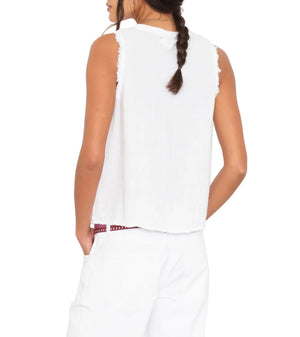 A woman with a braid is shown from the back wearing the Xirena Portia Top, a sleeveless white cotton gauze piece, paired with white pants and her left hand in her pocket.