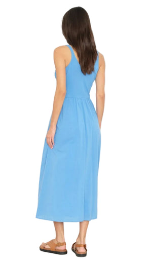 A woman stands with her back to the camera against a white background, wearing the Xirena Fallon Dress—a lightweight cotton poplin sleeveless midi dress with a full skirt—paired with brown sandals.