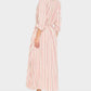 A woman stands sideways in the Xirena Bowen Stripe Dress, a long pink and white vertically striped shirt dress with rolled sleeves and a waist tie, crafted from a soft viscose-linen blend, styled with black and brown flat sandals against a white background.