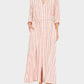 She wears the Xirena Bowen Stripe Dress—red and white vertical stripes, rolled sleeves, tie waist—paired with black sandals. This maxi A-line dress pops against the plain backdrop.