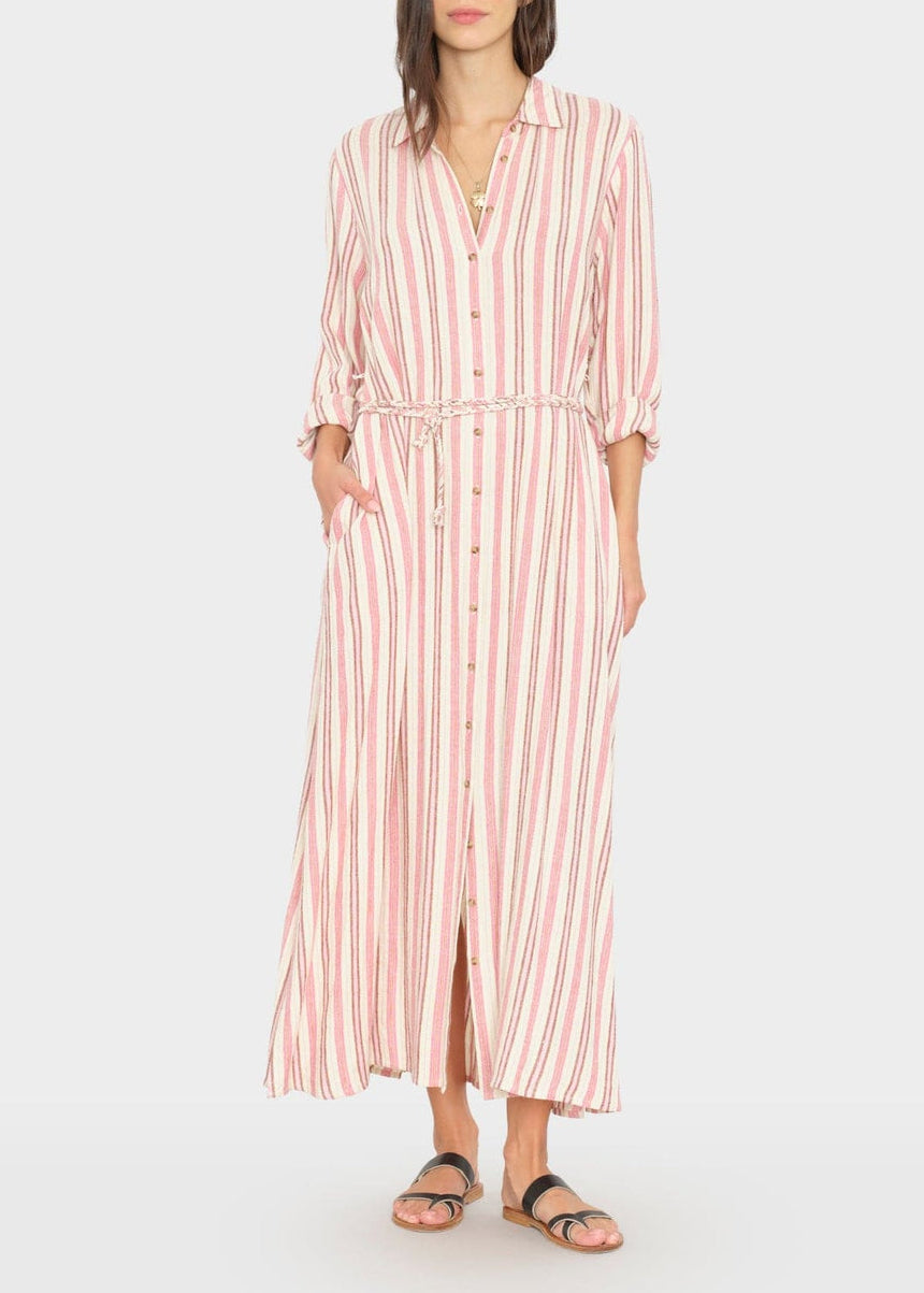 She wears the Xirena Bowen Stripe Dress—red and white vertical stripes, rolled sleeves, tie waist—paired with black sandals. This maxi A-line dress pops against the plain backdrop.