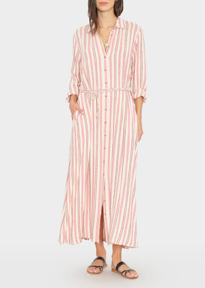 She wears the Xirena Bowen Stripe Dress—red and white vertical stripes, rolled sleeves, tie waist—paired with black sandals. This maxi A-line dress pops against the plain backdrop.