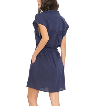 A woman is shown from the back with hands in pockets, wearing the Eliza Dress by Xirena—a navy blue cotton poplin mini shirt dress with cap sleeves—against a white background.