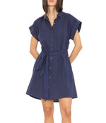 A woman wears the Xirena Eliza Dress, a navy blue cotton poplin mini shirt dress with a tied waist belt, standing against a plain white background.