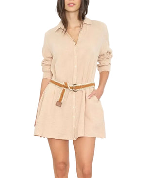 A woman wears the Xirena Bobby Cotton Gauze Dress in light beige, featuring long sleeves, a braided belt, and a relaxed fit with one hand casually tucked in her pocket.