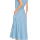 A woman stands with her back turned, wearing the Xirena Dixie Chambray Dress—a light blue, short-sleeve, button-front cotton midi dress—paired with leopard print sandals.